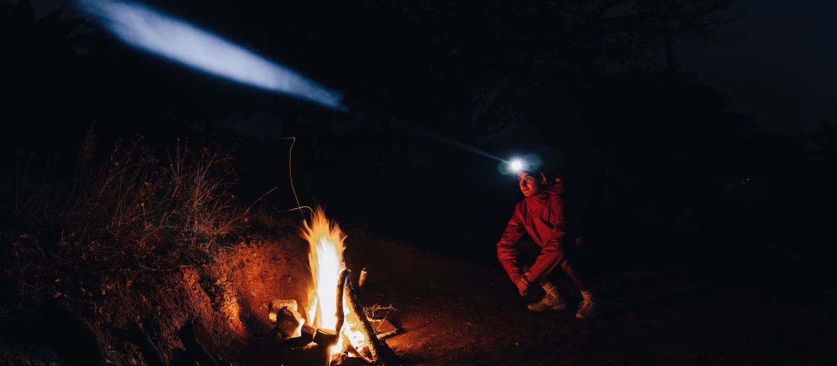 Man with headlamp sitting by campfire at night, surrounded by nature.