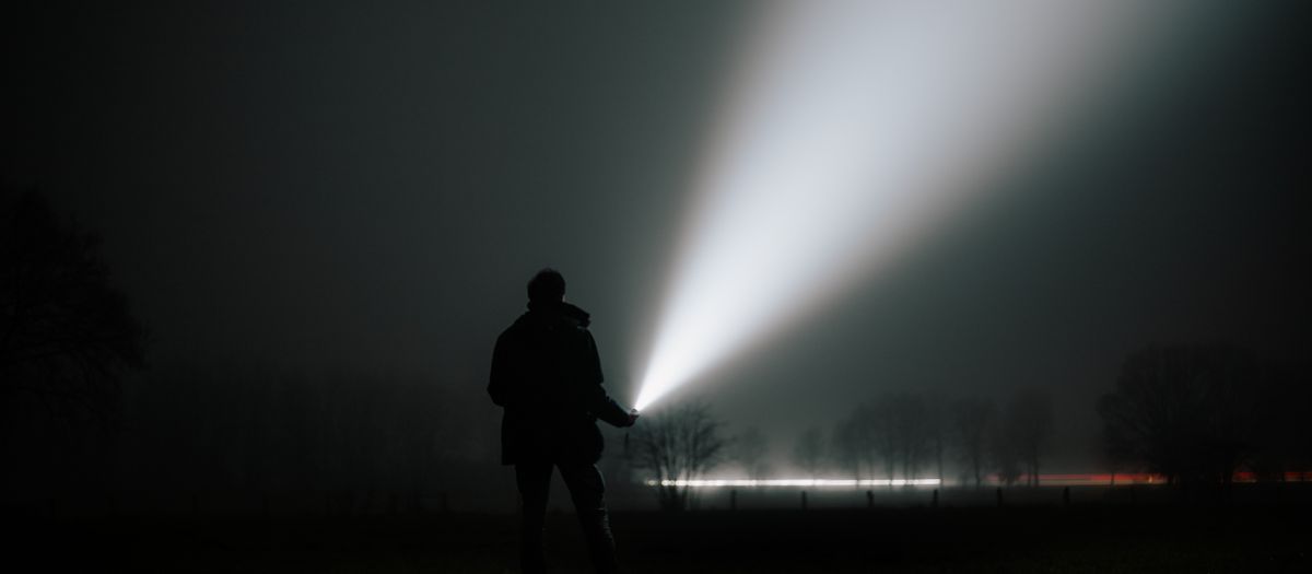A person holds a flashlight in a foggy night landscape, creating dramatic light trails.