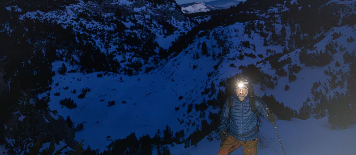 A solitary hiker climbs snowy mountains at night, guided by a headlamp.
