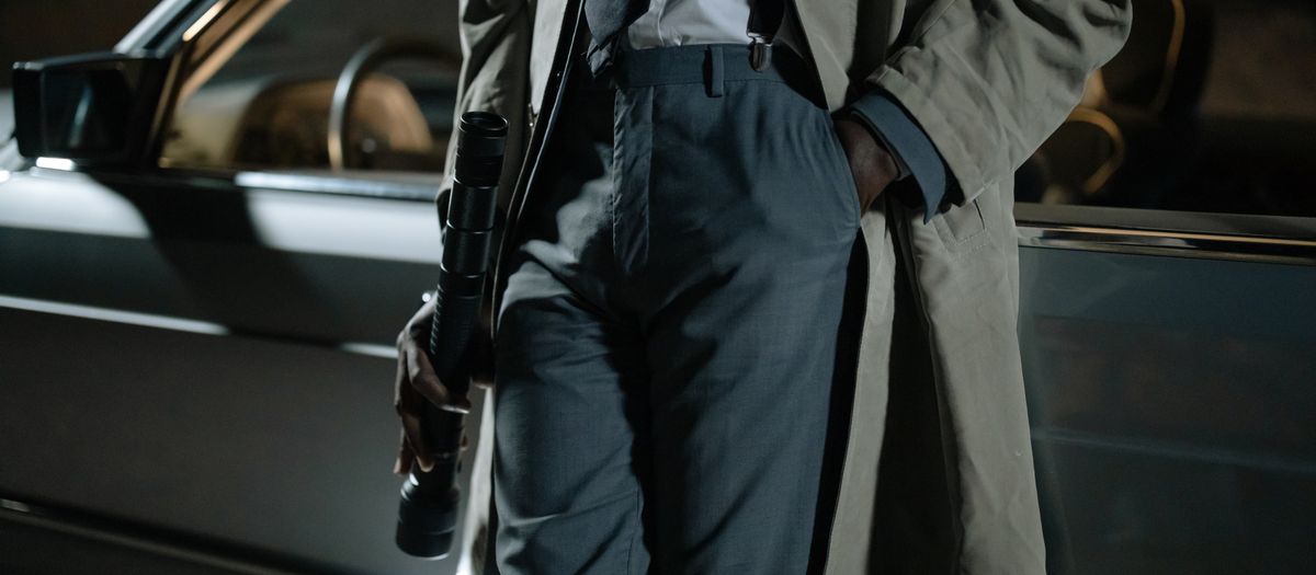 Close-up of a person in a trench coat holding a flashlight near a car at night.