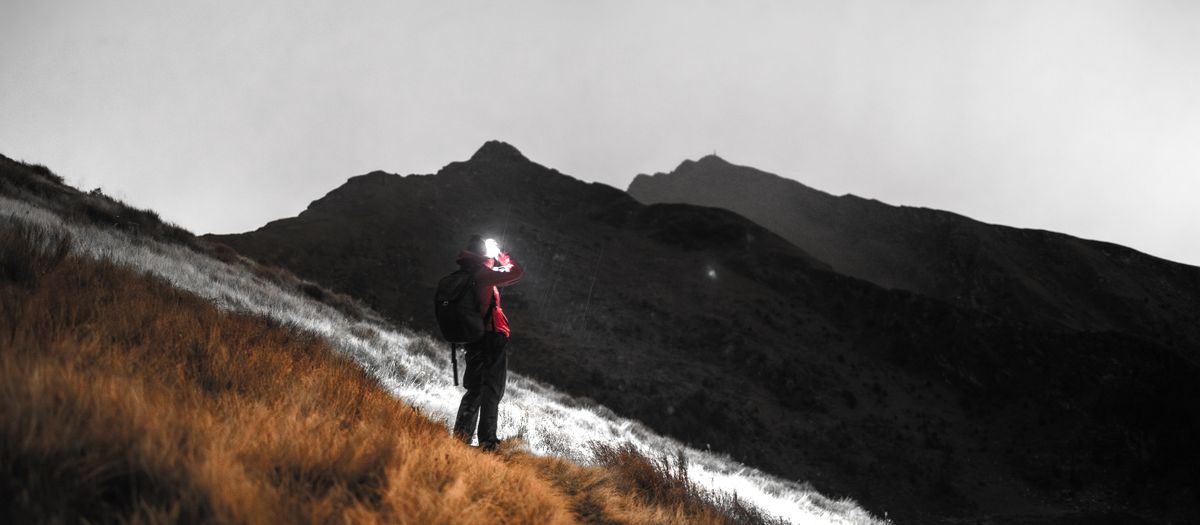 A lone hiker illuminated by a headlamp traverses a mountain path under a cloudy sky.