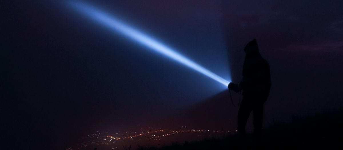 A person holds a flashlight, casting a beam at night with city lights in the background in Virgínia, Brazil.